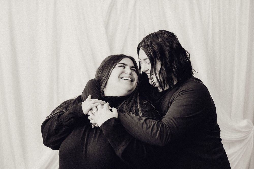 Two young women laugh and embrace playfully in a black and white studio portrait.