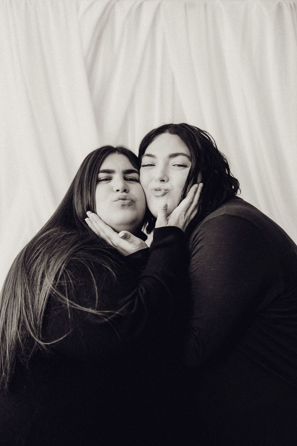 Two young women in black tops make kissing faces in a playful black and white portrait.