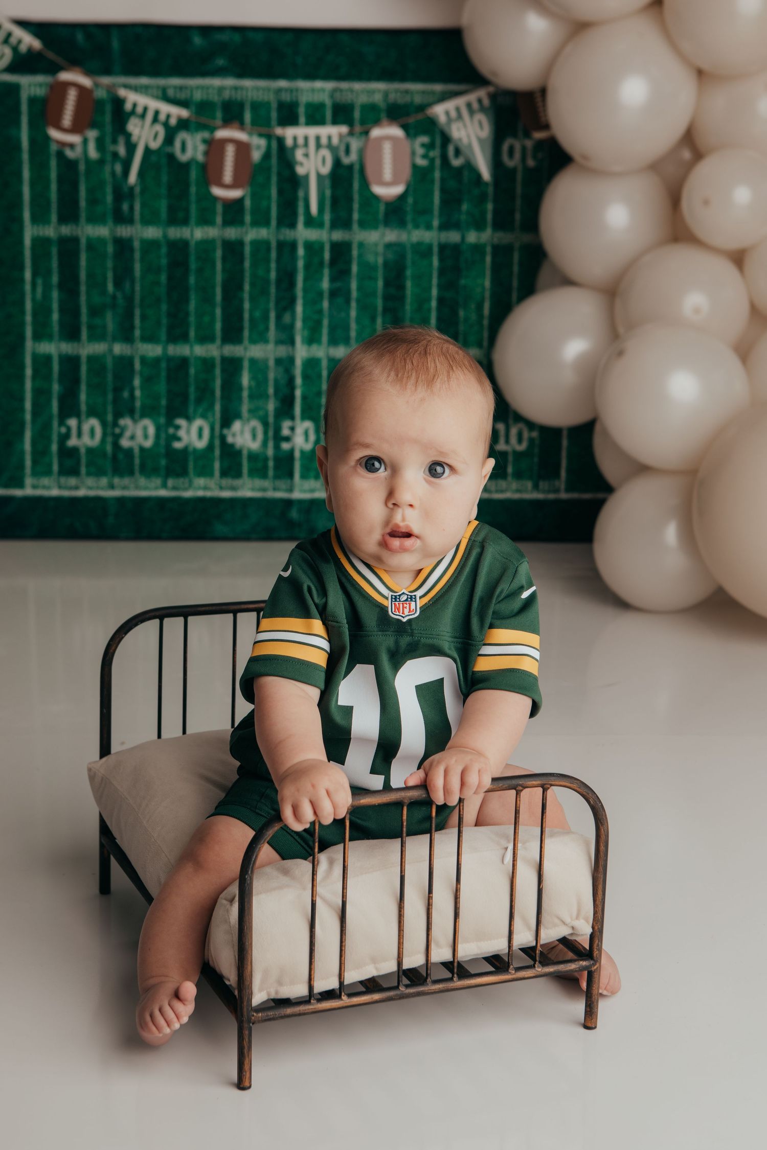 Football-themed baby photo shoot with green field backdrop and white balloons.