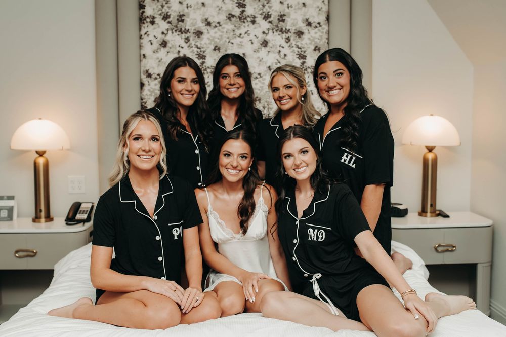 Bride in white with bridesmaids wearing matching black monogrammed pajamas posing on a bed.