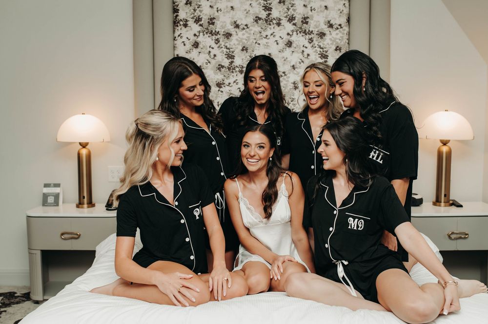 Bride and bridesmaids in black monogrammed pajamas laughing together on a hotel bed.