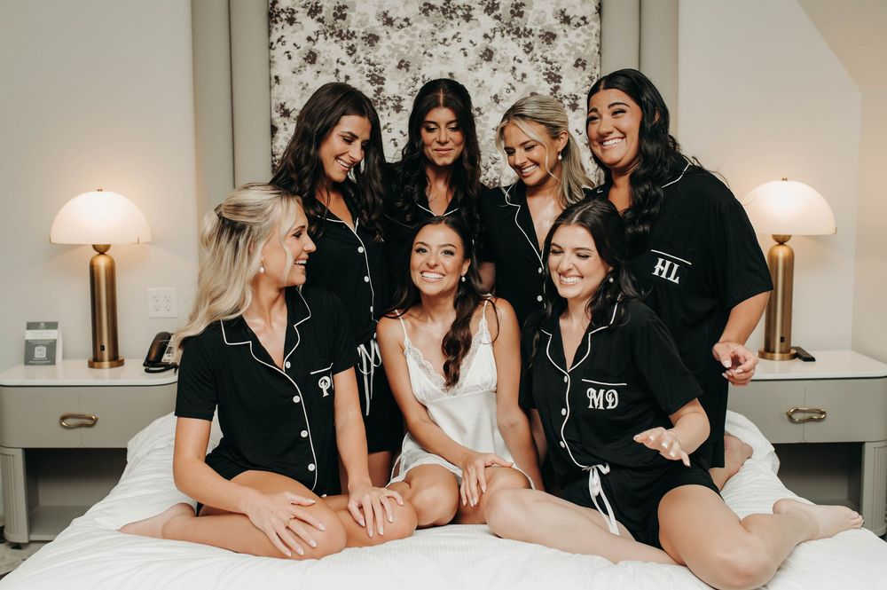 Bride in white surrounded by smiling bridesmaids in black monogrammed pajamas on a bed.