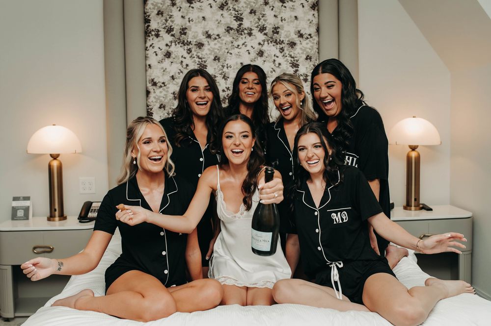 Bride popping champagne with excited bridesmaids in black pajamas celebrating on a hotel bed.