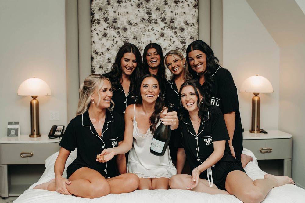 Bridesmaids in matching black monogram pajamas celebrate with champagne on a white bed.