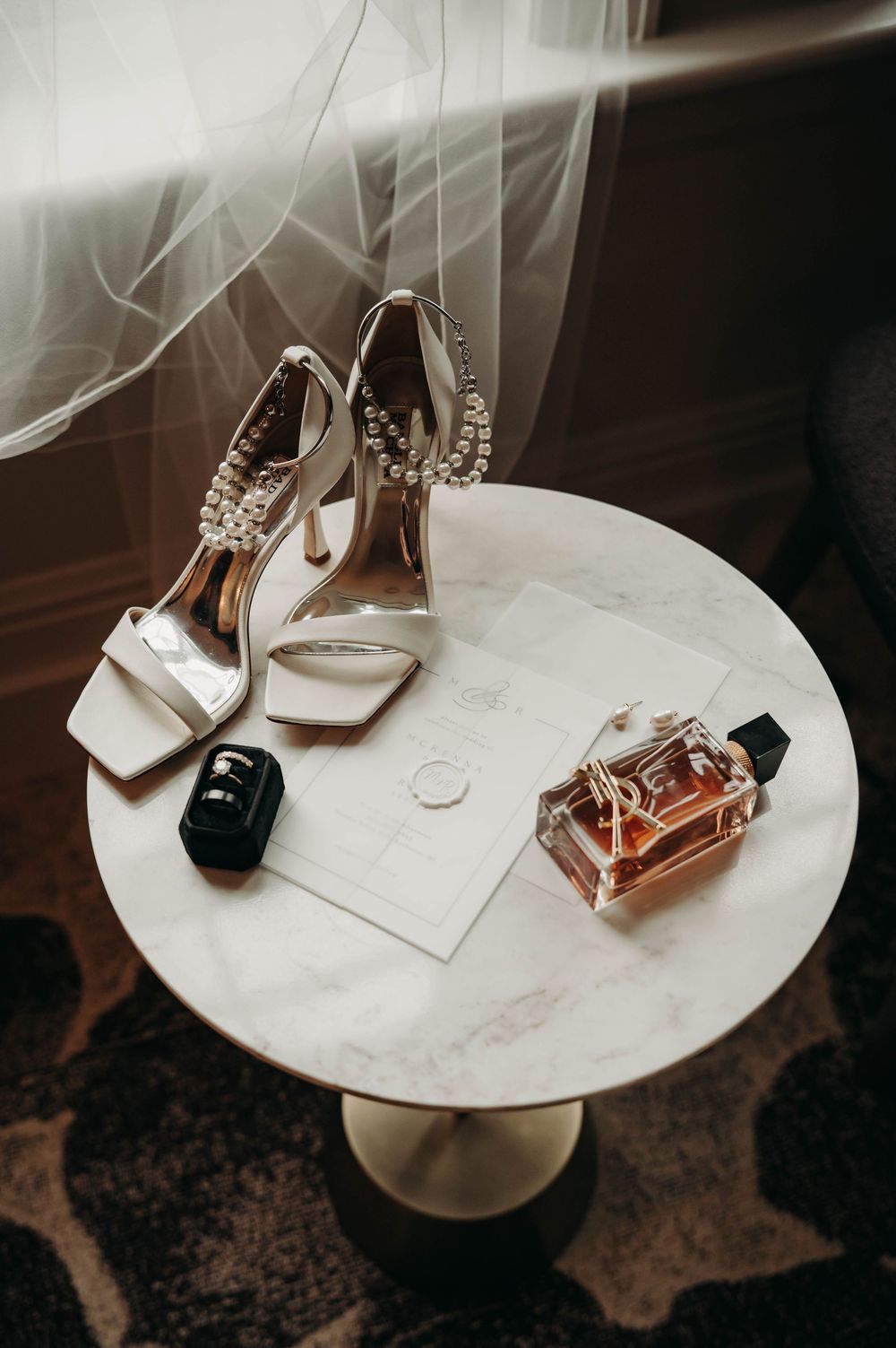 Bridal flat lay featuring pearl-adorned heels, wedding rings, perfume bottle, and invitation on marble table.