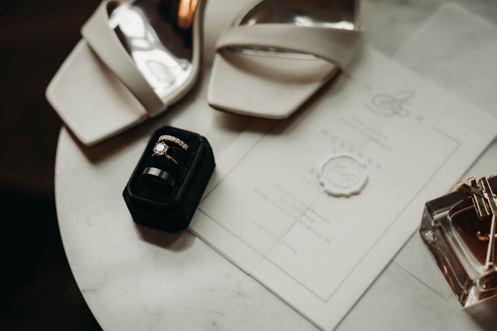 Close-up of wedding rings in black velvet box beside bridal heels, perfume, and wax-sealed invitation.