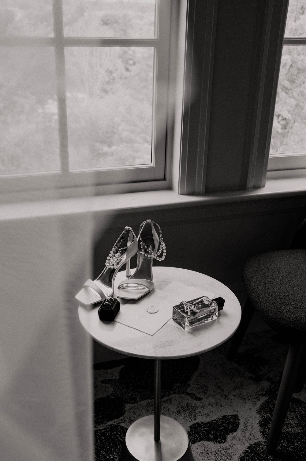 Black and white image of bridal accessories including heels, rings, and perfume on marble table by window.