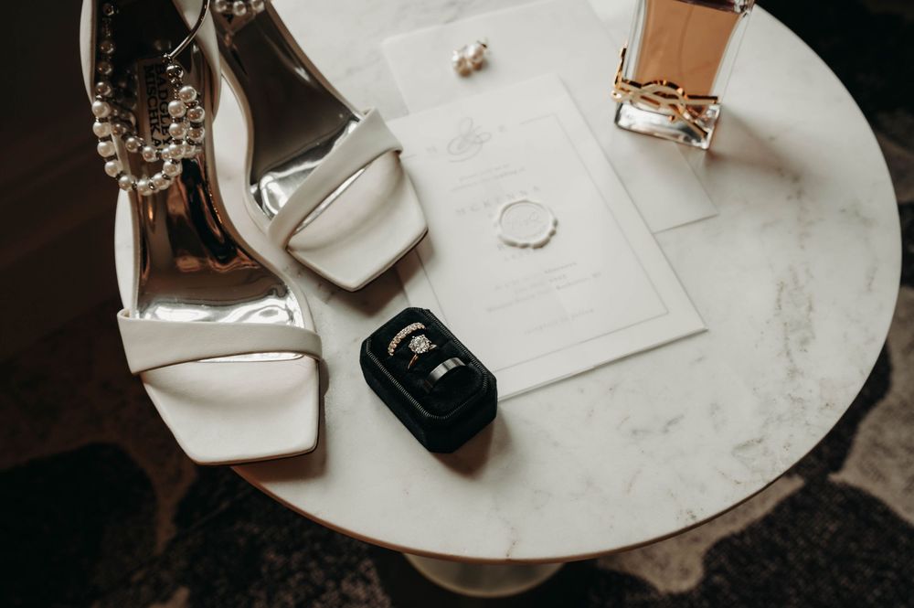 White bridal heels, wedding rings, invitation, and perfume arranged on a marble table.