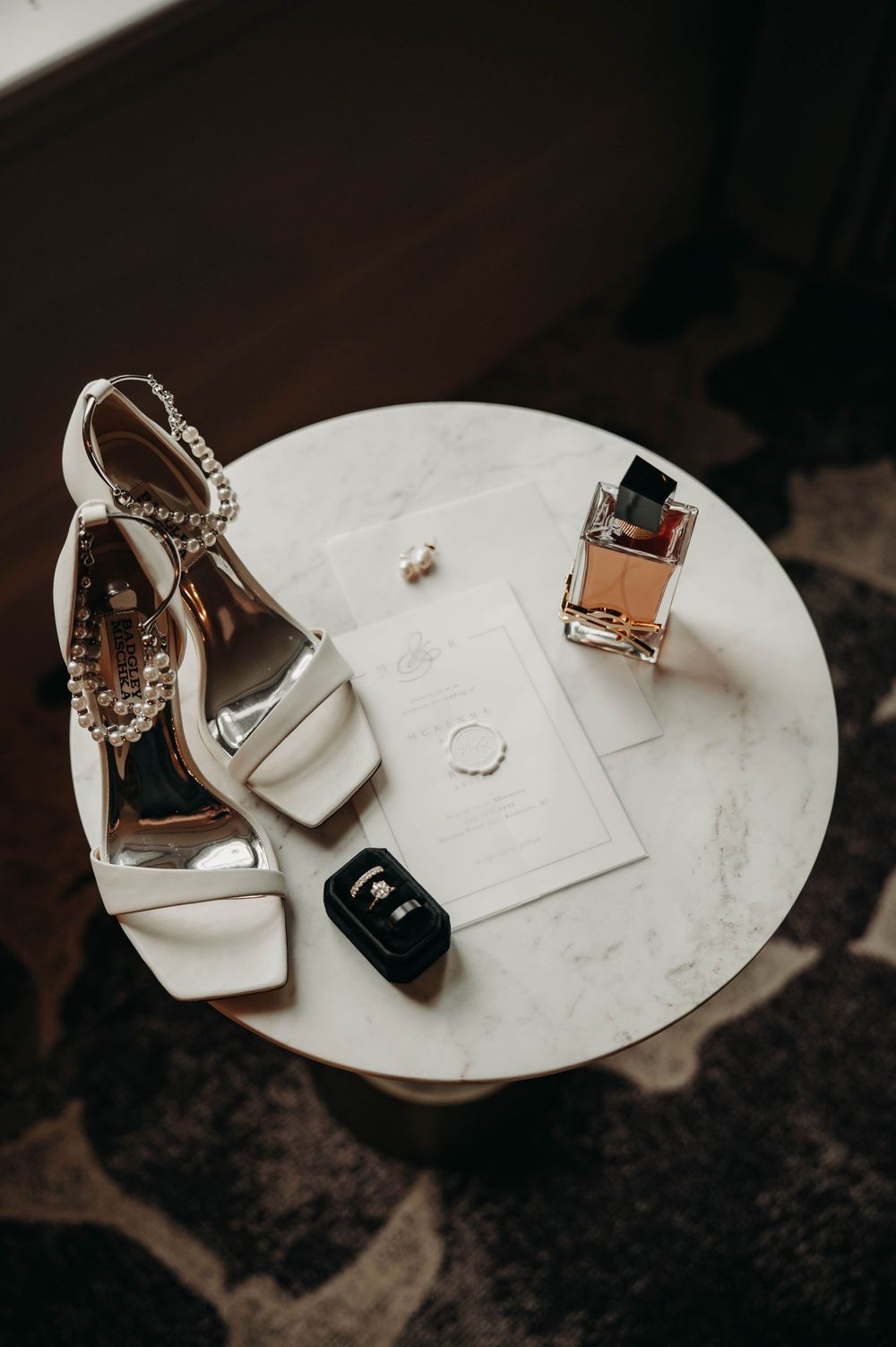 Elegant bridal flat lay with white heels, perfume, rings, and wedding invitation on marble.