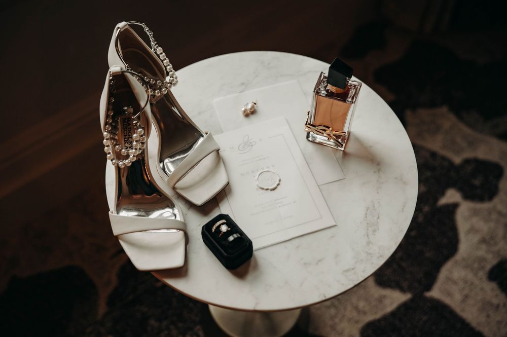 Bridal accessories including white heels, perfume bottle, and ring box on a marble table.