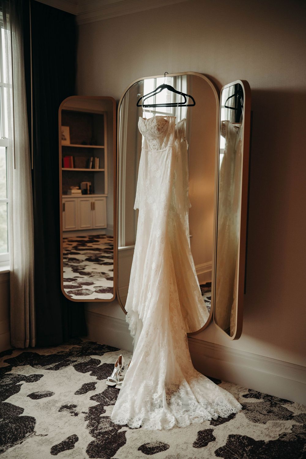 Lace wedding dress hanging on a gold tri-fold mirror in a softly lit hotel room.