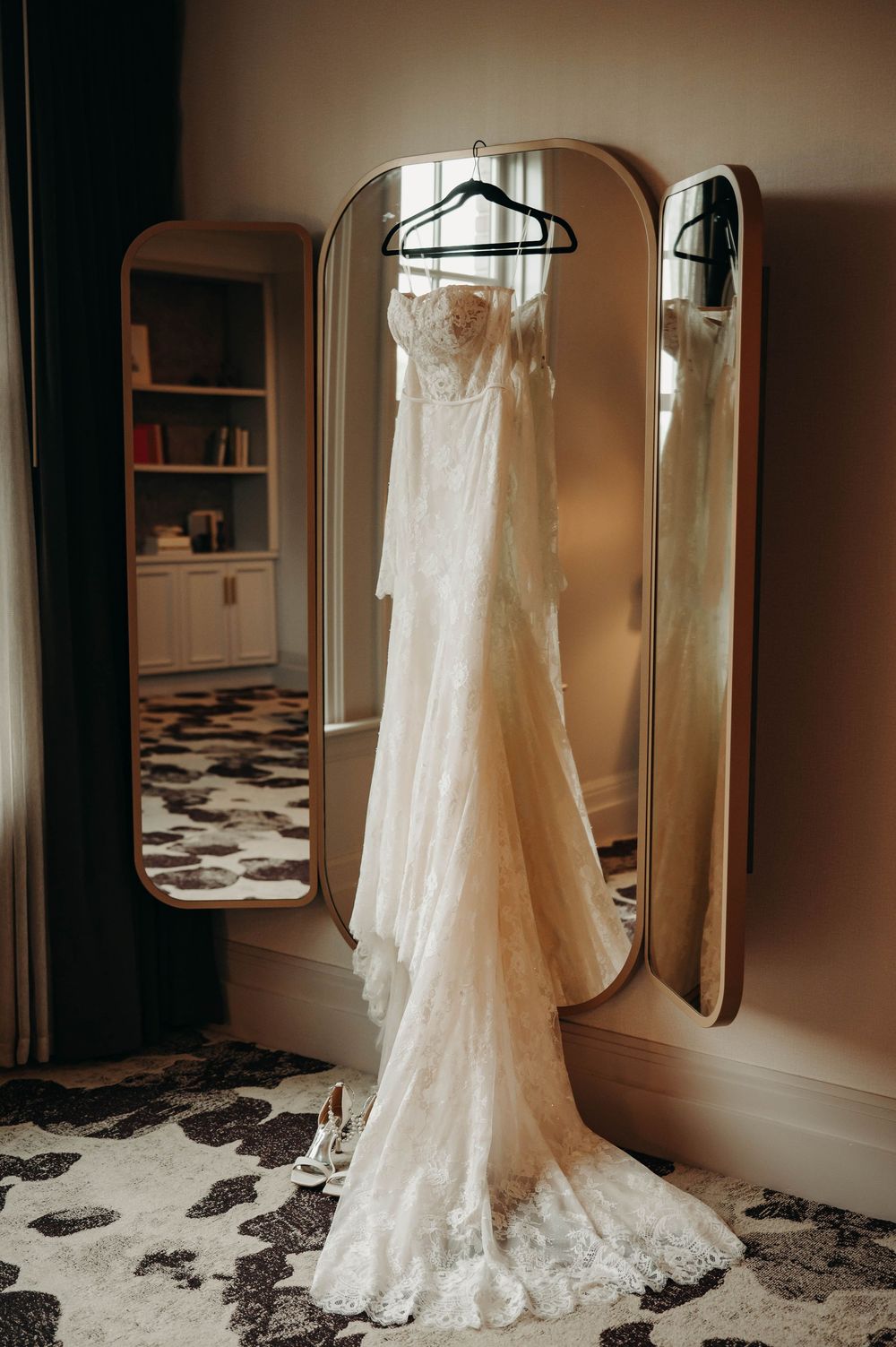 Elegant lace bridal gown displayed on a hanger over a gold trifold mirror indoors.
