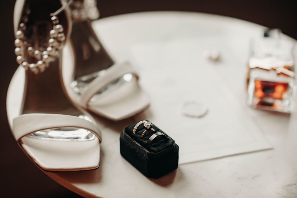 Wedding rings in black box displayed beside white heeled bridal shoes and perfume bottle on table.