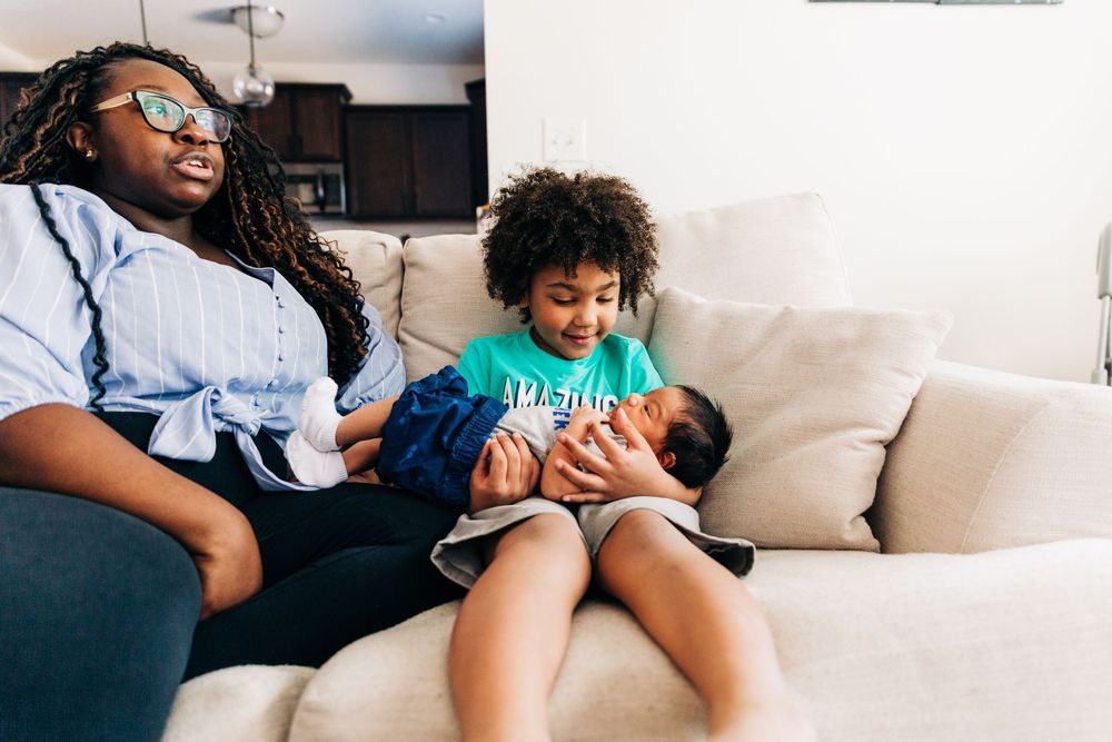A mother sits on a beige couch next to her young son who is holding a newborn baby.