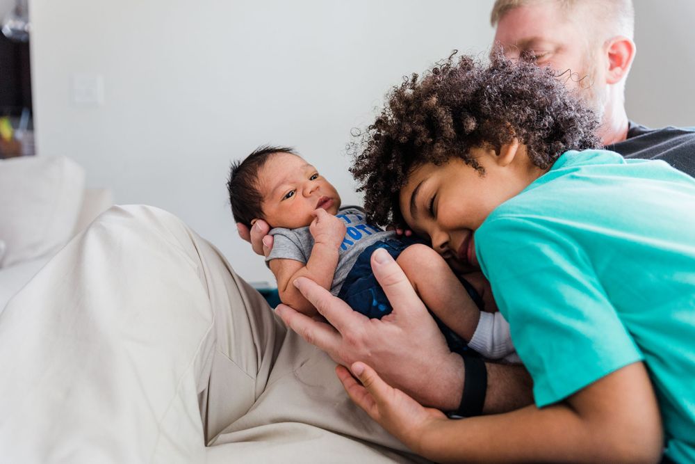 A big brother cuddles his newborn brother while dad holds the baby.