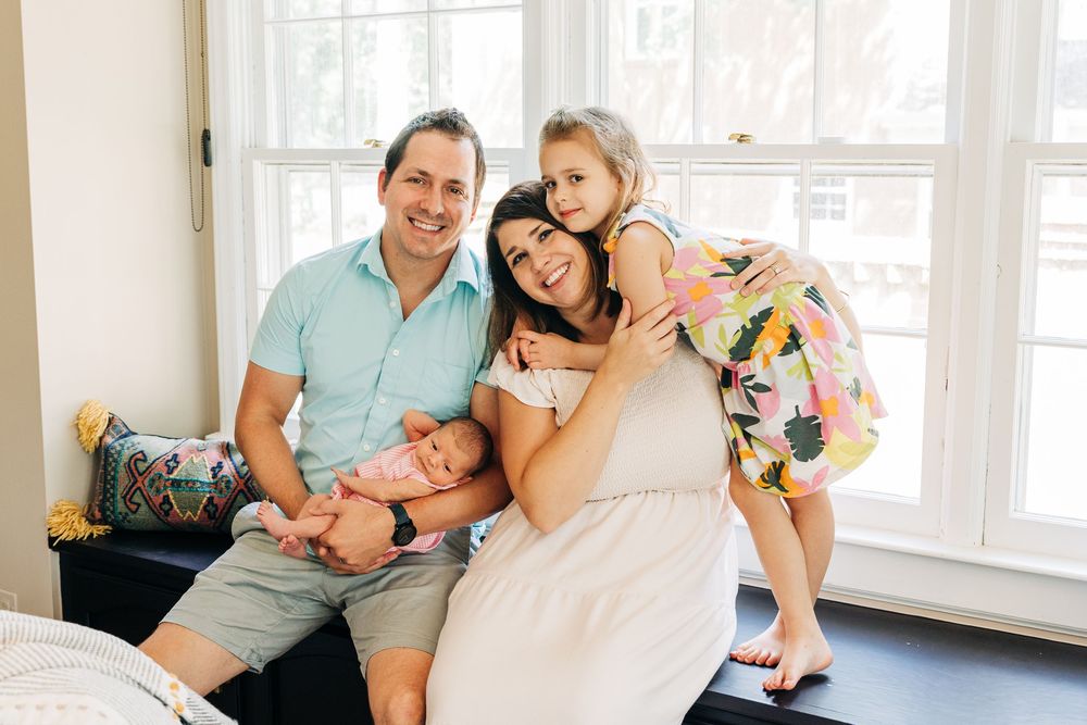 A family relaxes and cuddles together on a couch in a bright, sunlit living room for newborn photos.
