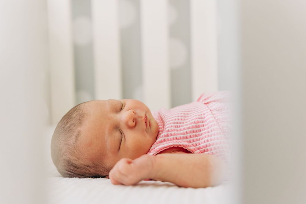 A newborn baby sleeps peacefully in a white crib wearing a pink and white striped outfit during Charlotte newborn photo session