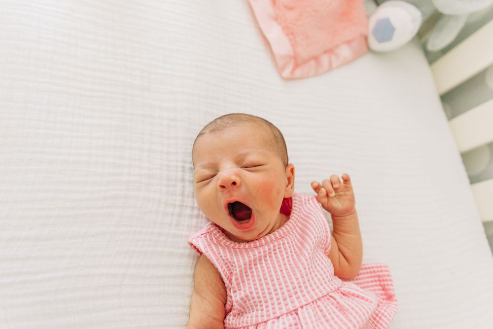 A newborn baby in a white crib wearing a pink gingham outfit stretches and yawns during Charlotte NC photo session.