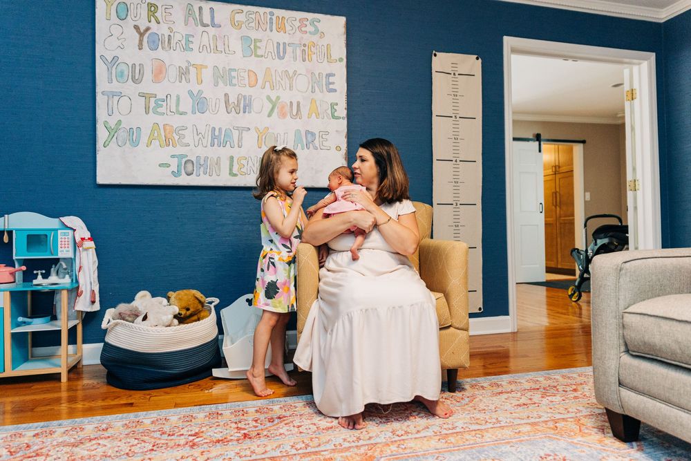 A mother holding her newborn in the playroom with her preschooler. Blue walls and toys are all around.