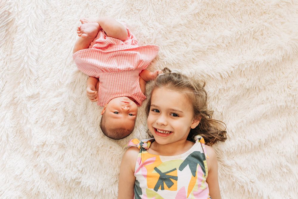 A young child lies on white bedding while smiling next to her newborn baby sister during Charlotte photo session.