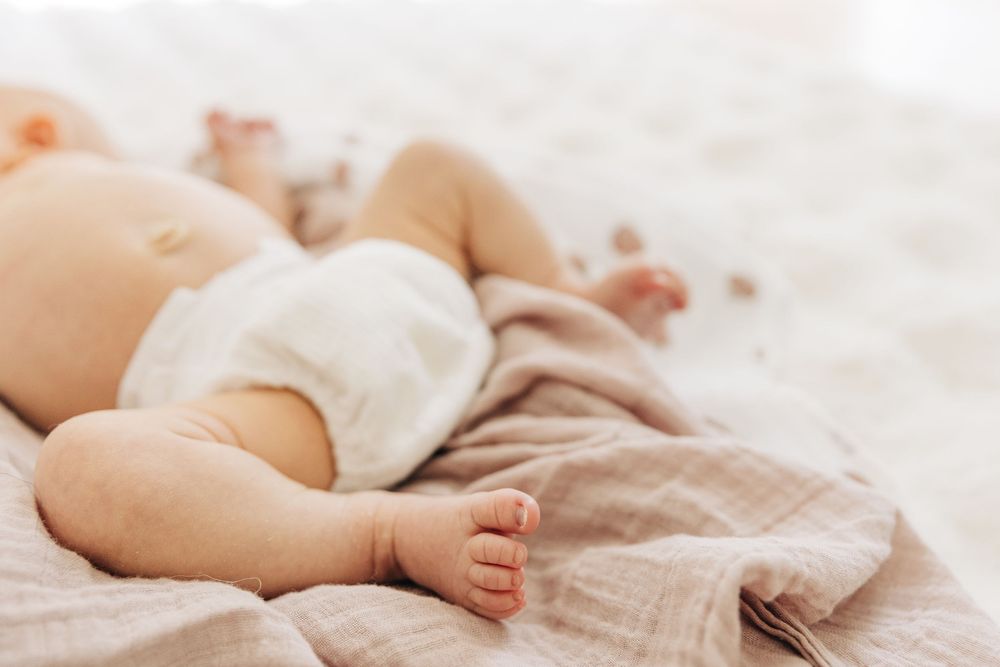 A sleeping baby in a soft beige outfit lies on white bedding with tiny feet peeking out.