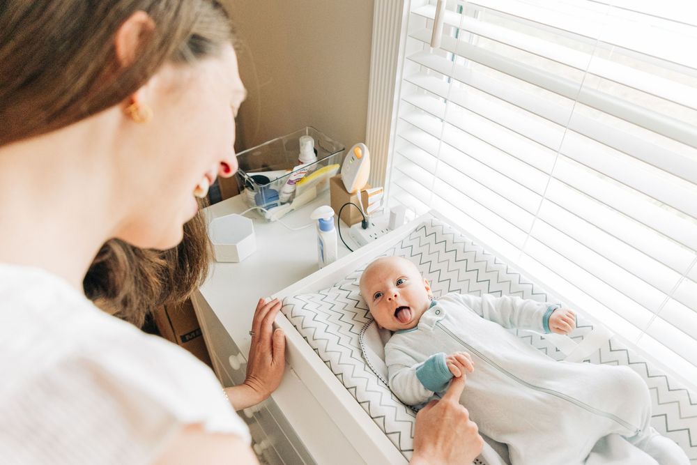 A newborn baby smiles up at his mom while lying on a grey changing pad.