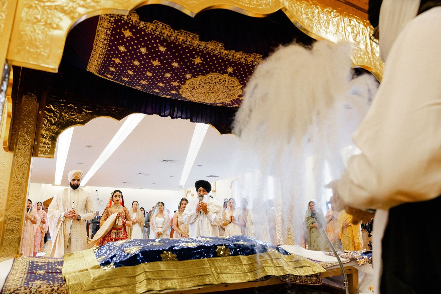 Wide shot of a Sikh wedding ceremony (Anand Karaj) inside a Hounslow Gurdwara, capturing the couple and family in prayer