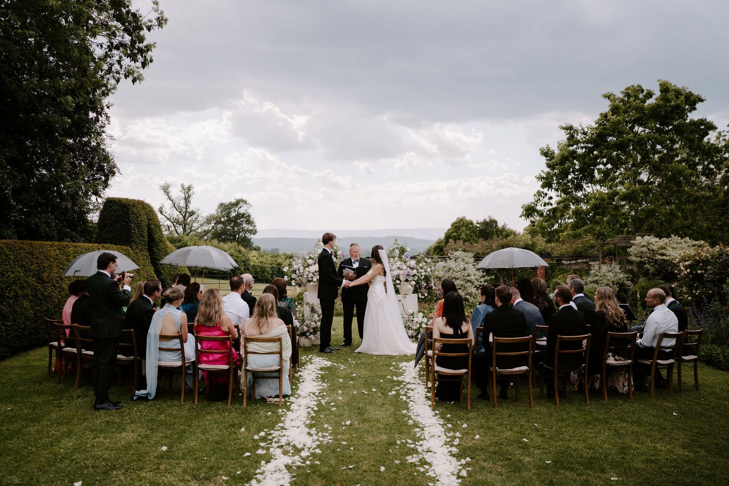 wedding guests, bride and groom sheltering underneath umbrellas during a rain shower during the ceremony at dewsall court in herefordshire