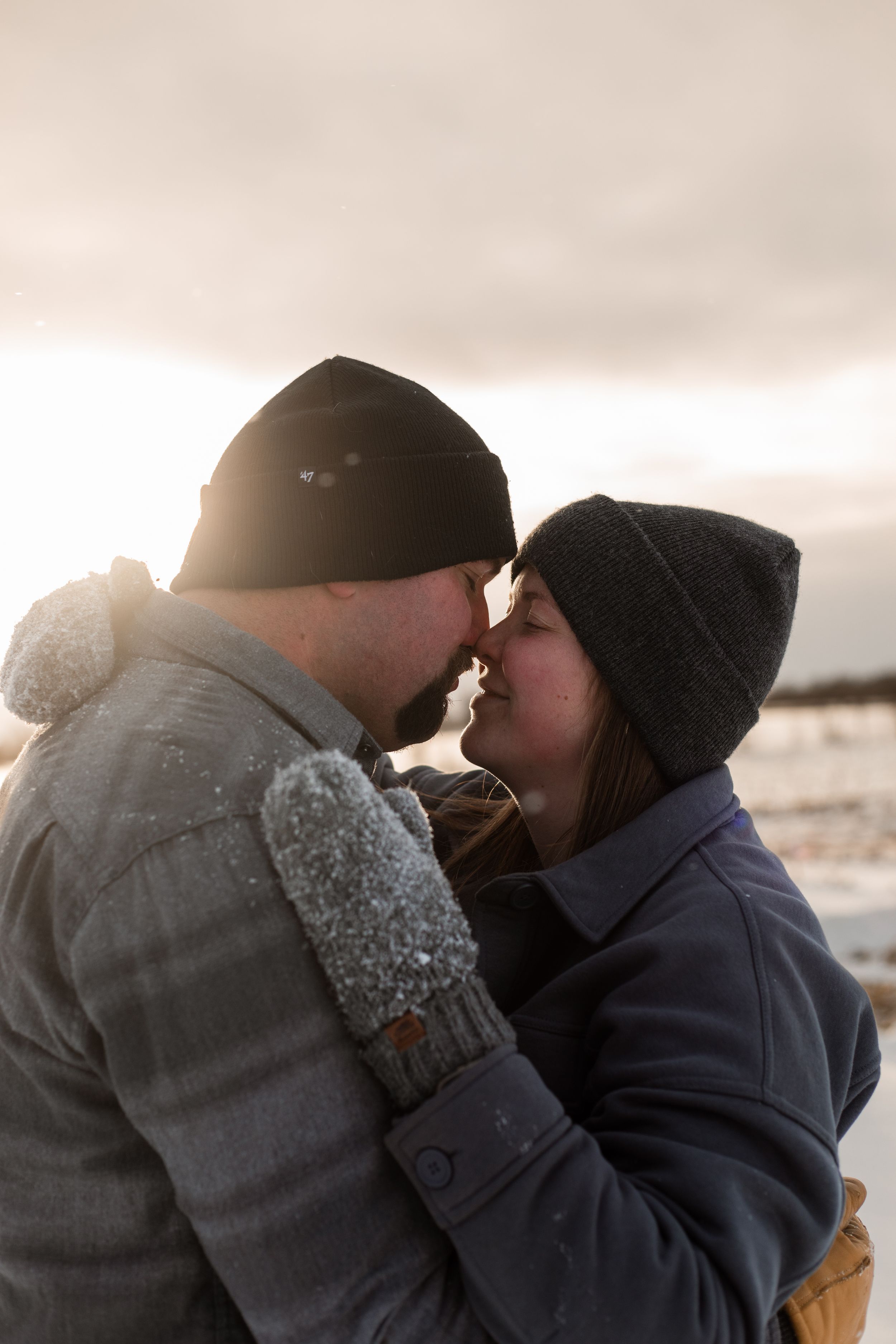 A couple in winter clothing share an intimate moment at sunset against a cloudy sky.