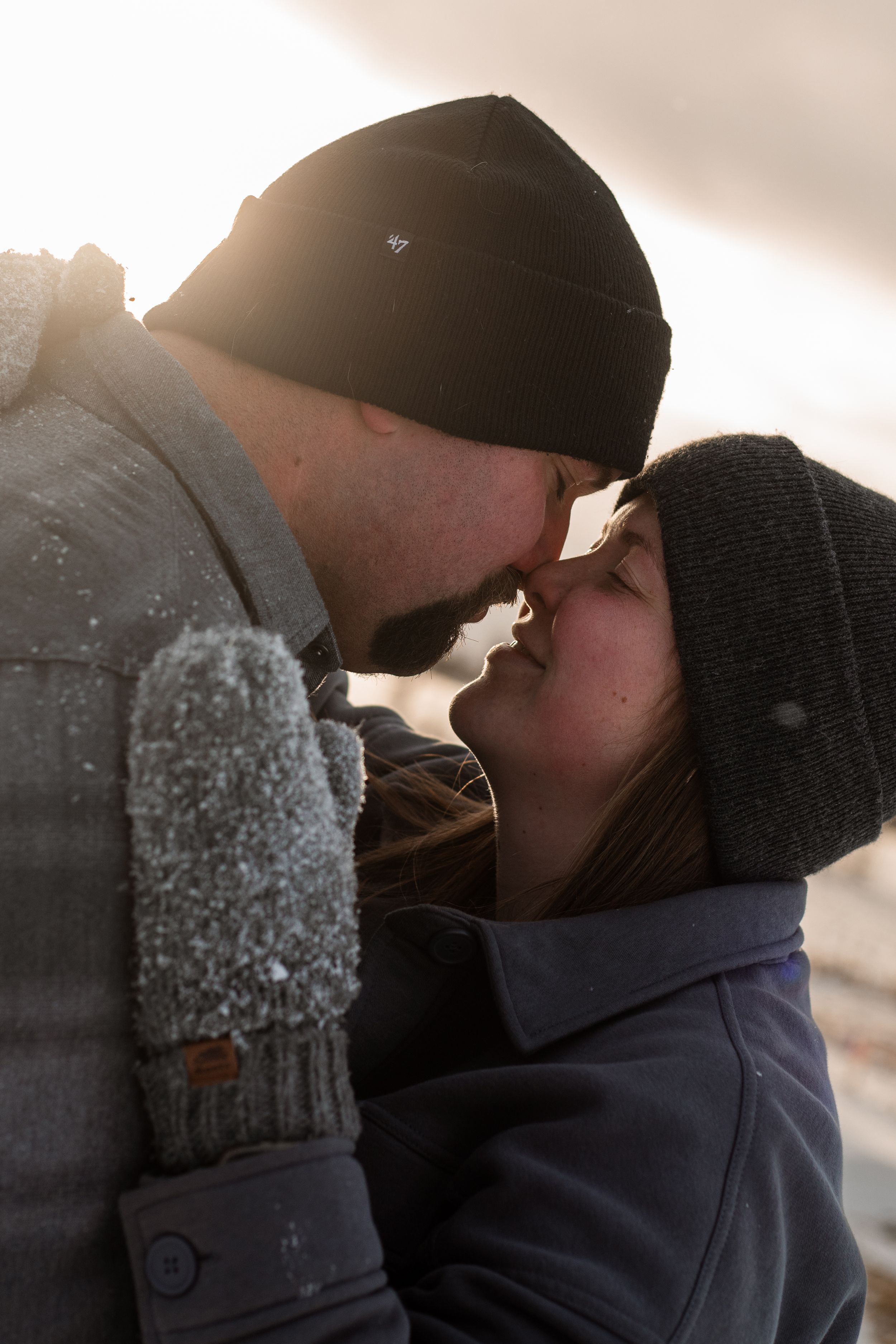 A romantic winter scene shows a couple in knit hats sharing an intimate moment at sunset with snowflakes falling around them.
