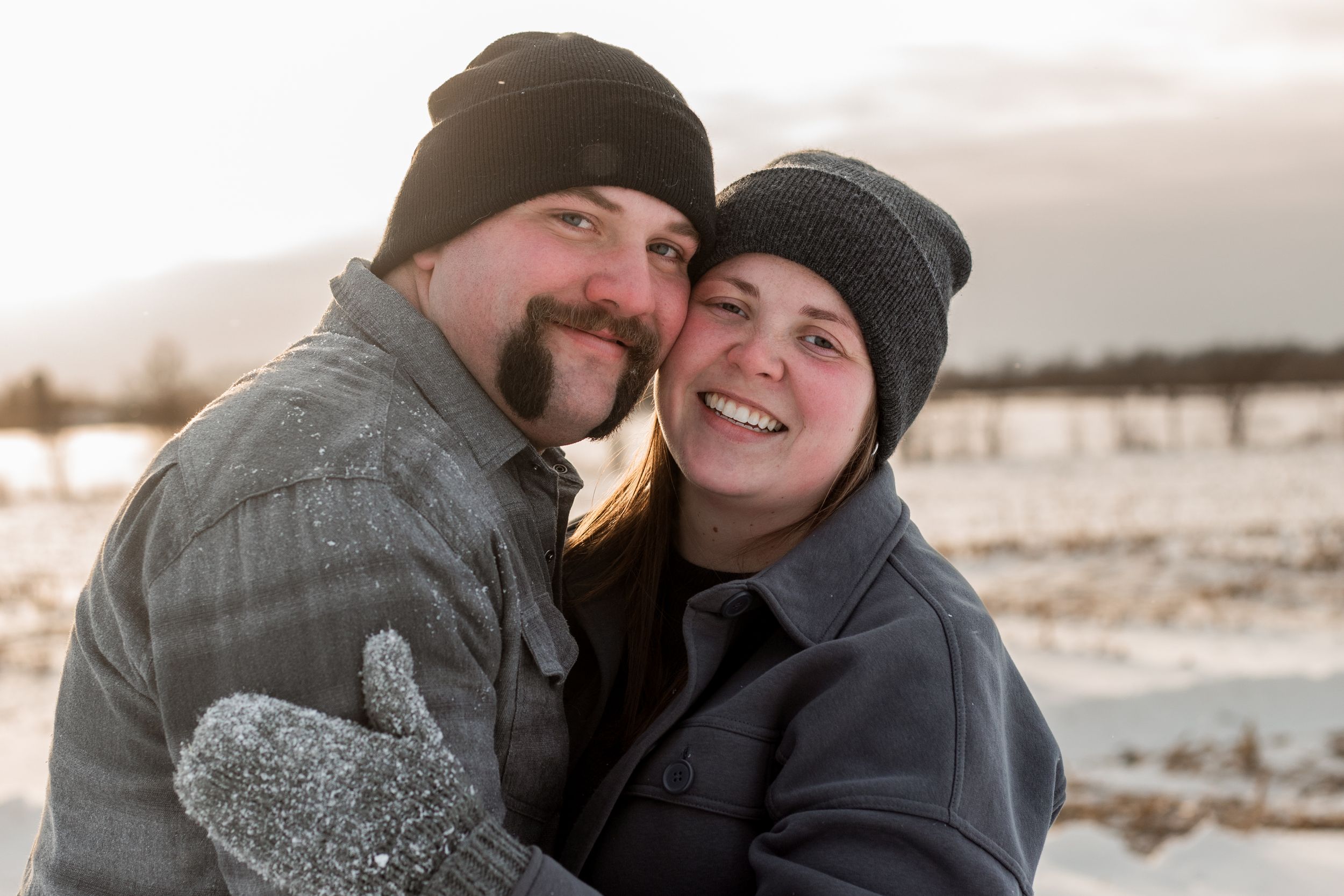 A couple wearing winter hats embraces and smiles while enjoying a snowy winter landscape at sunset.