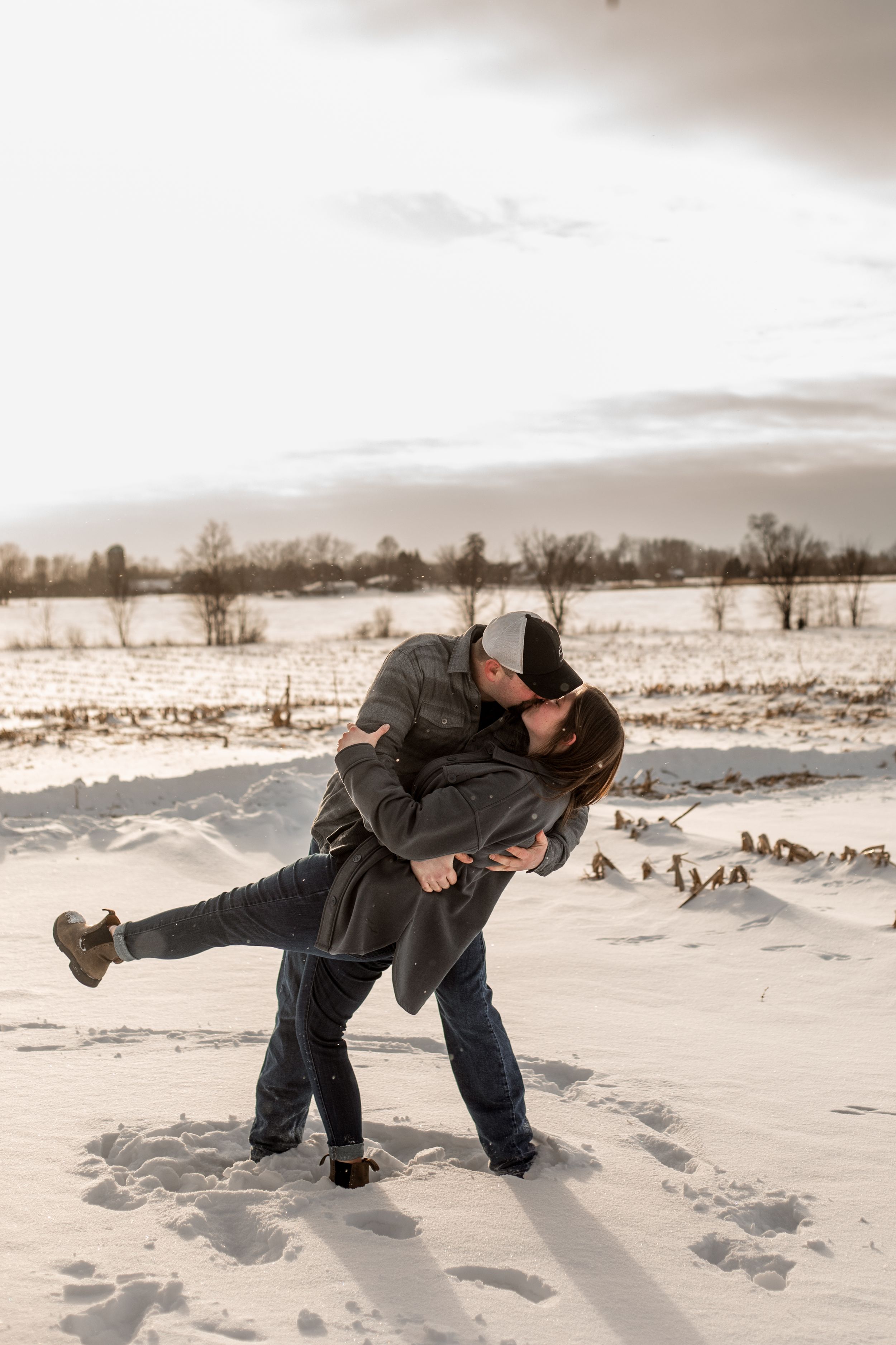 A romantic winter scene shows a couple embracing in a dramatic dip pose against a snowy rural landscape at dusk.