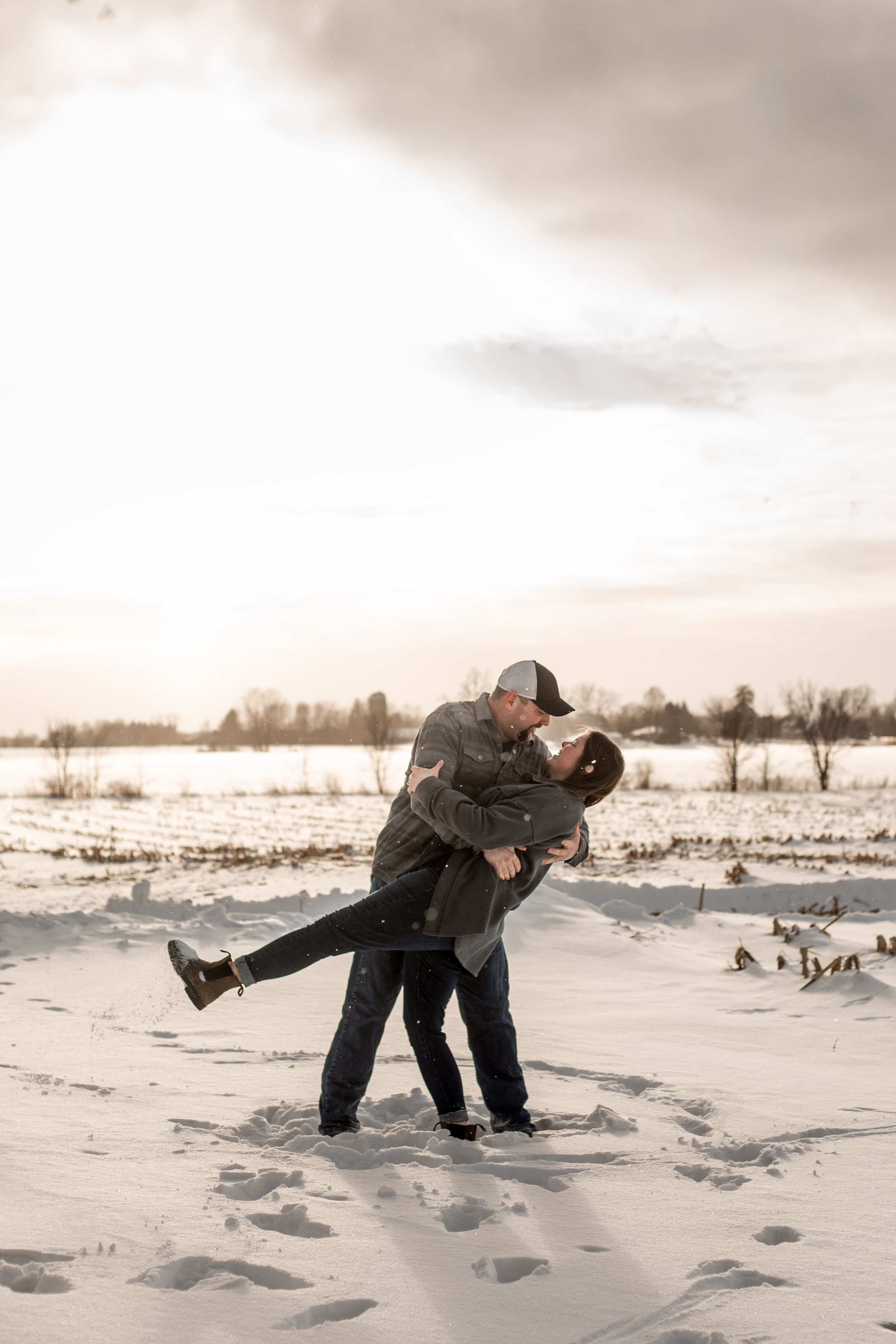 A couple embraces in a romantic winter scene with snow-covered ground and geese in the background at sunset.