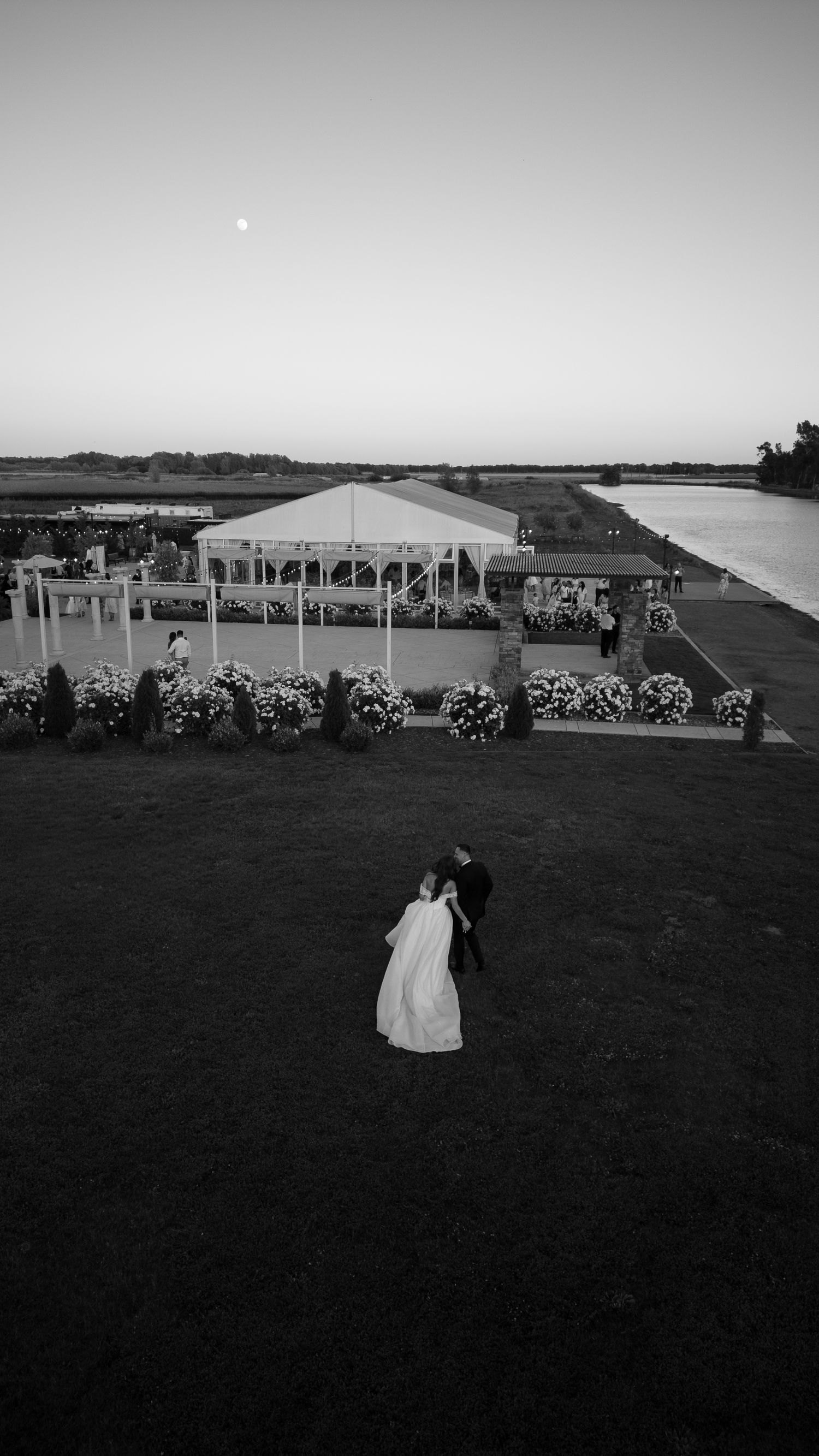 A wedding reception setup at dusk with white tents and chairs arranged on a grassy lawn overlooking a waterfront view.