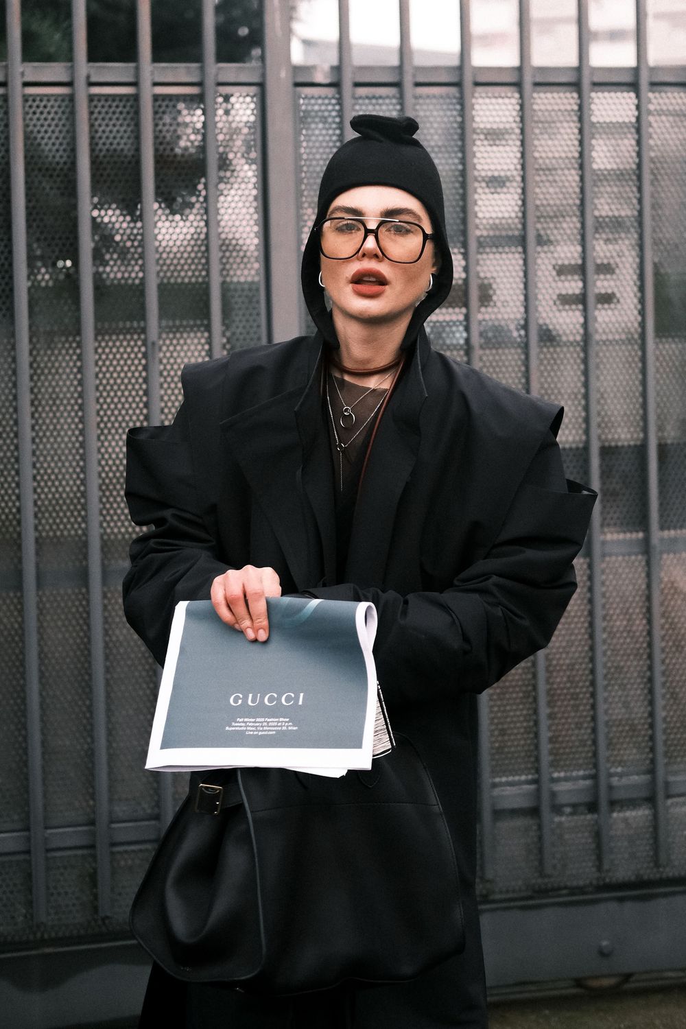 A person in dark clothing and glasses poses against an industrial fence backdrop with a dramatic aesthetic.