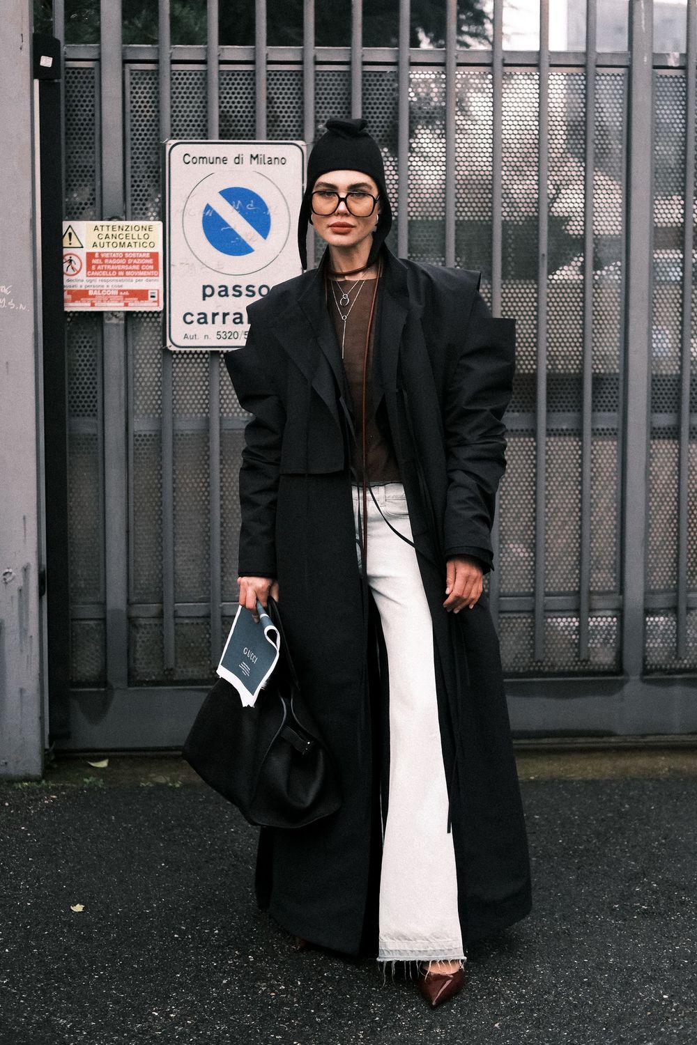 Two people dressed in black coats and white dresses stand against a gray industrial fence in an urban setting.
