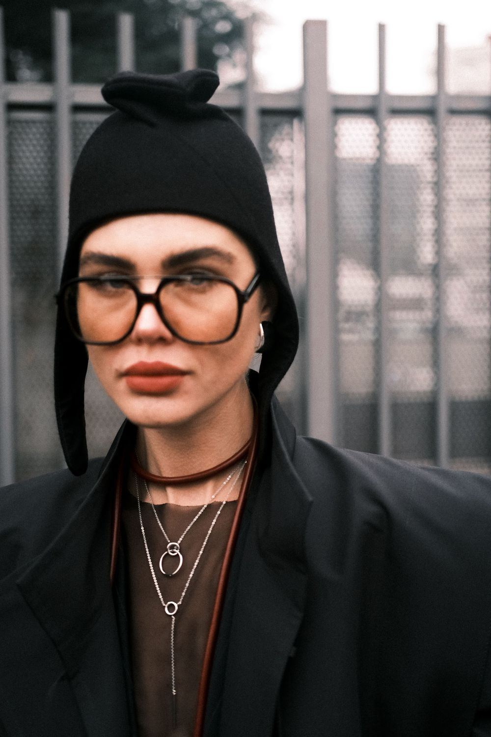 Portrait of someone wearing a black beanie, oversized glasses, layered necklaces and red lipstick against a blurred fence.