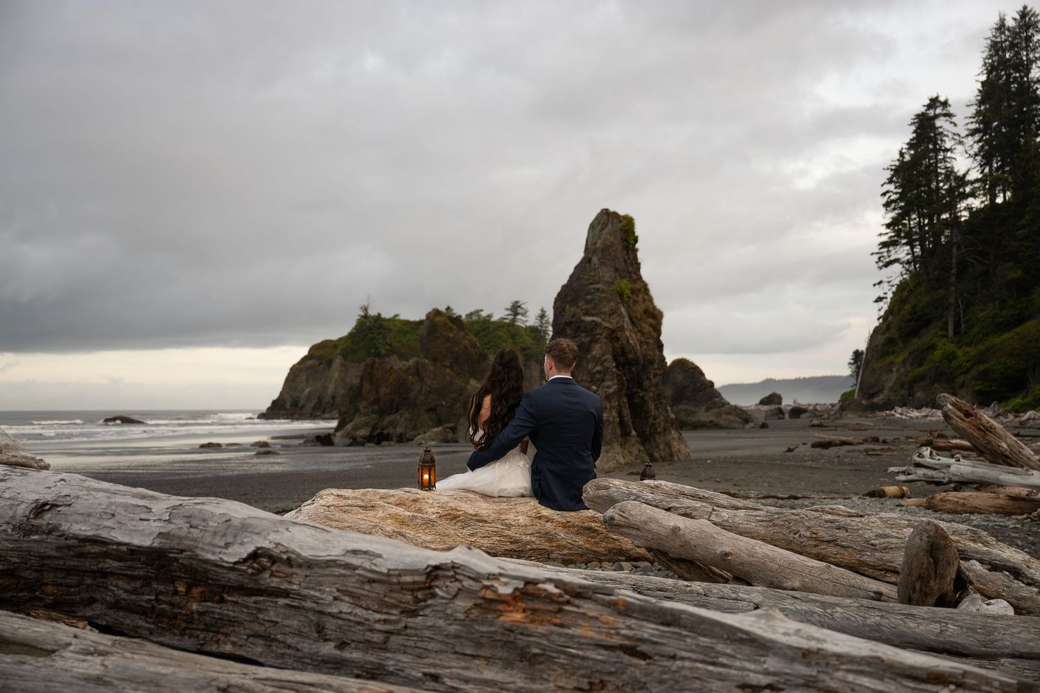 Newlyweds sit on driftwood with lanterns on an overcast day at Ruby Beach.