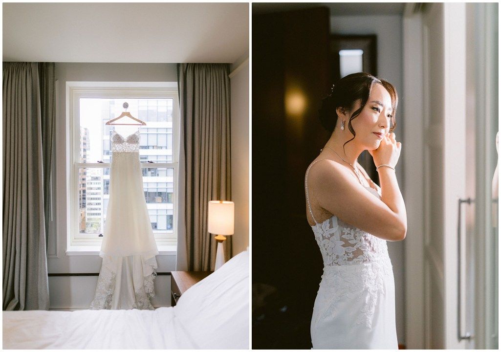 Elegant white lace wedding dress hangs by window as bride in matching gown stands pensively in soft natural light.