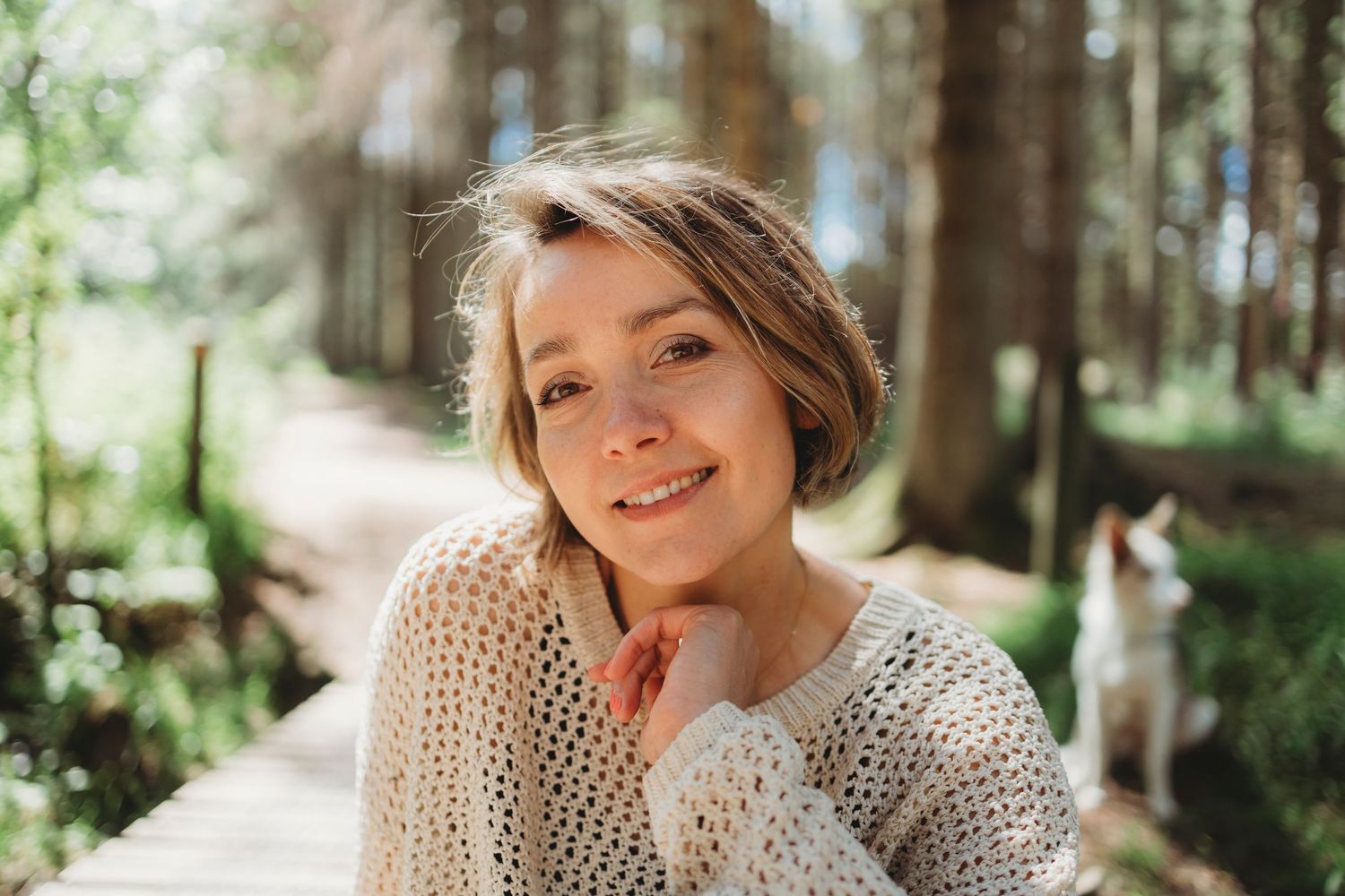 A person wearing a cream knit sweater poses in a sunlit forest setting with tall trees in the background.