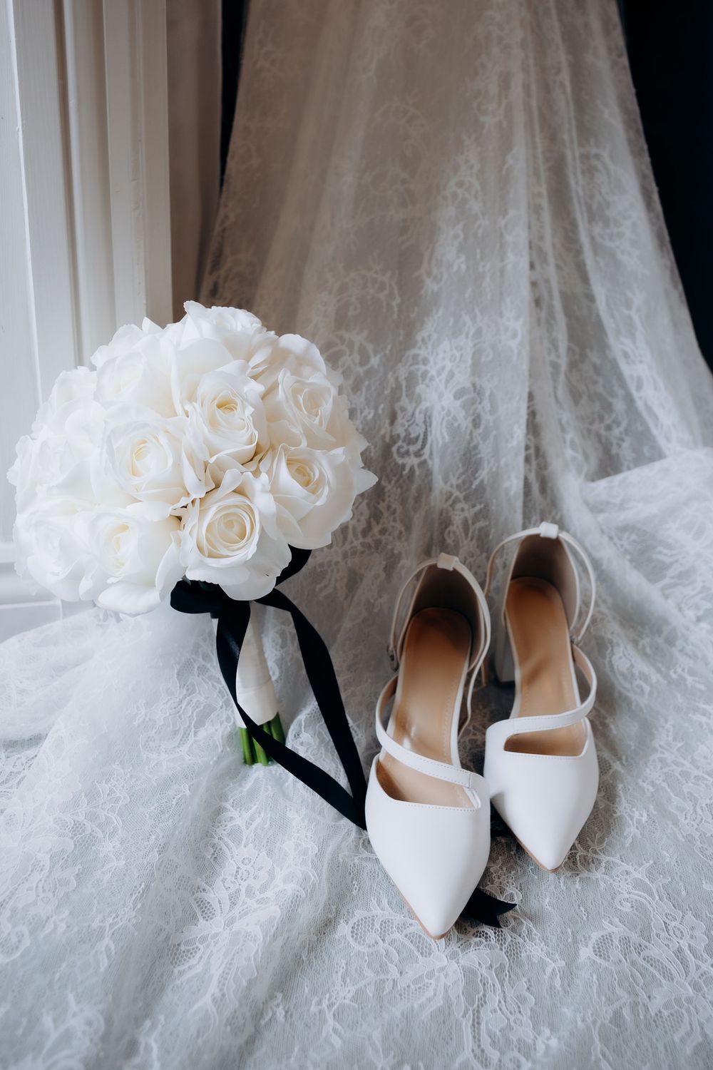 White bridal bouquet of roses and elegant white wedding shoes arranged on delicate lace fabric.