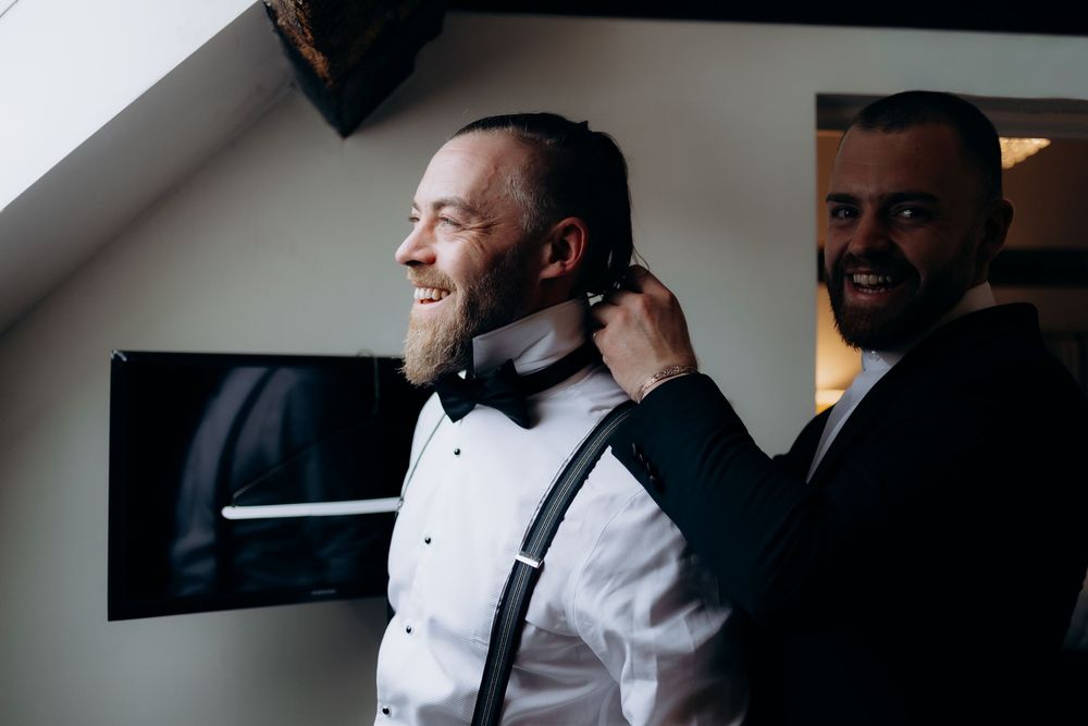 A person adjusts a black bowtie while wearing a white dress shirt and tuxedo jacket in a formal setting.