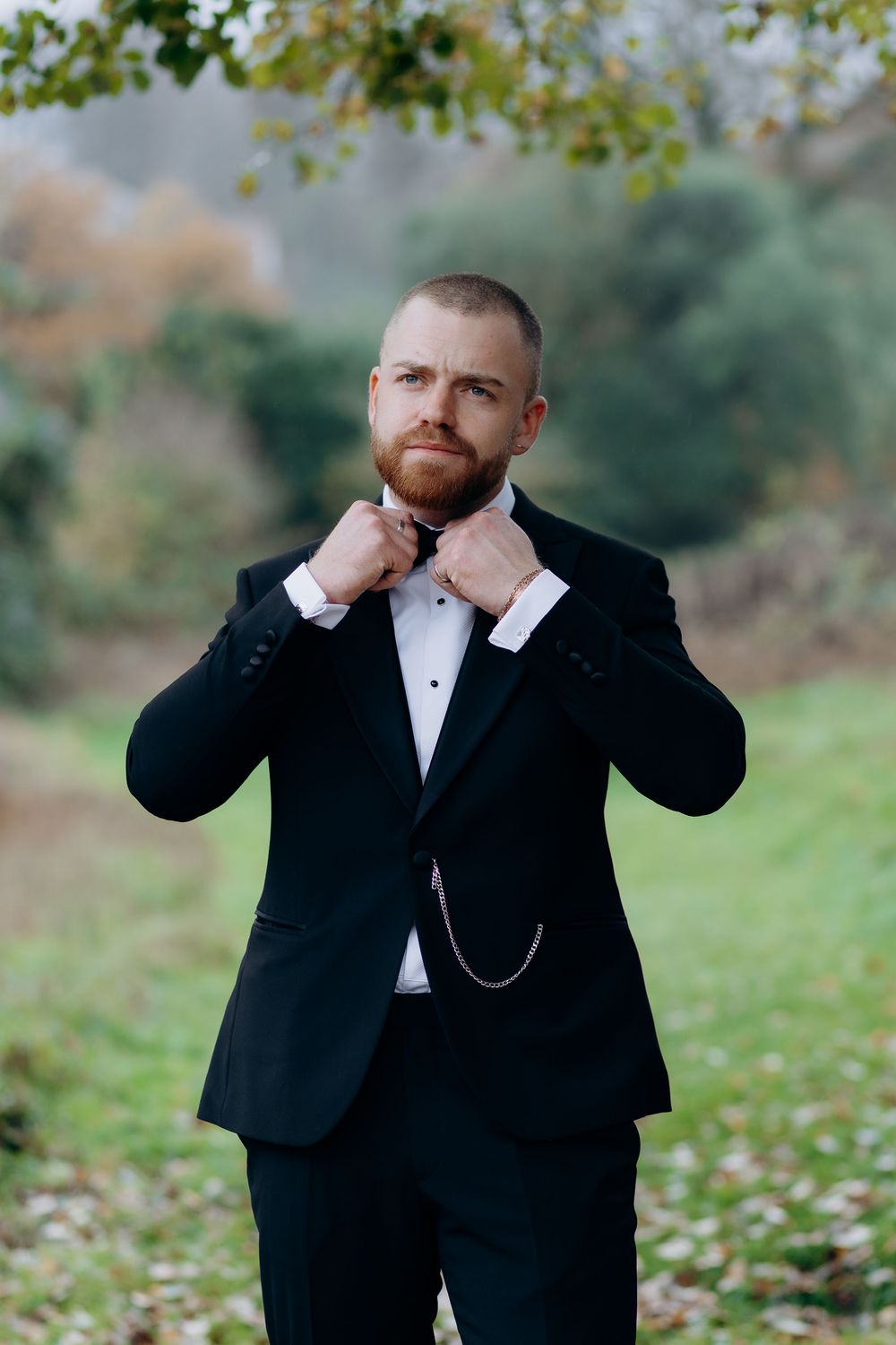 A groom in a black tuxedo poses outdoors in autumn while adjusting his bowtie and cuffs.
