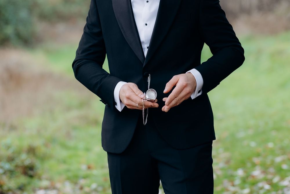 A groom in a black tuxedo adjusts his bow tie while standing outdoors on a grassy lawn.