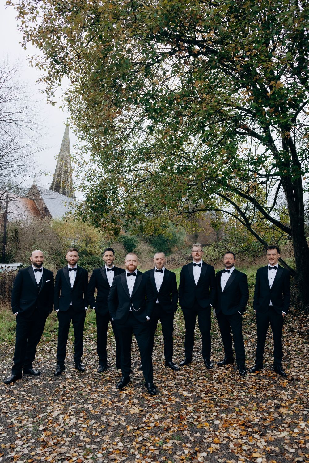 Groomsmen in black tuxedos stand in a line on fallen autumn leaves with trees and church steeple in background.