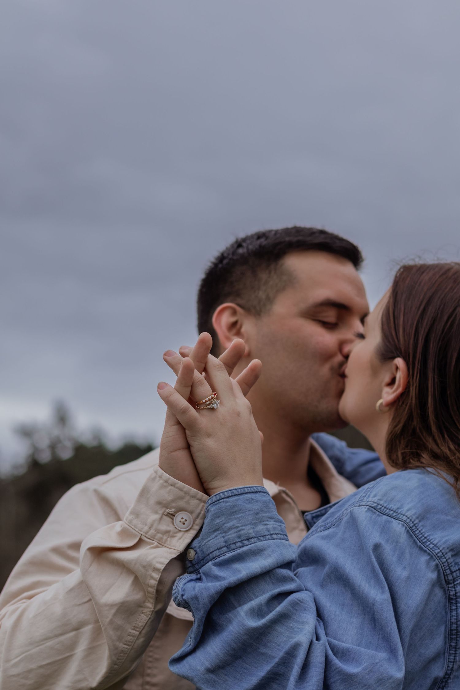 A couple shares a kiss outdoors under a cloudy sky, hands intertwined showing an engagement ring.