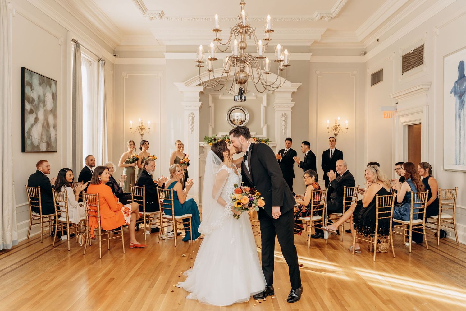 Elegant wedding ceremony in a bright white ballroom with crystal chandelier and guests seated on gold chairs.