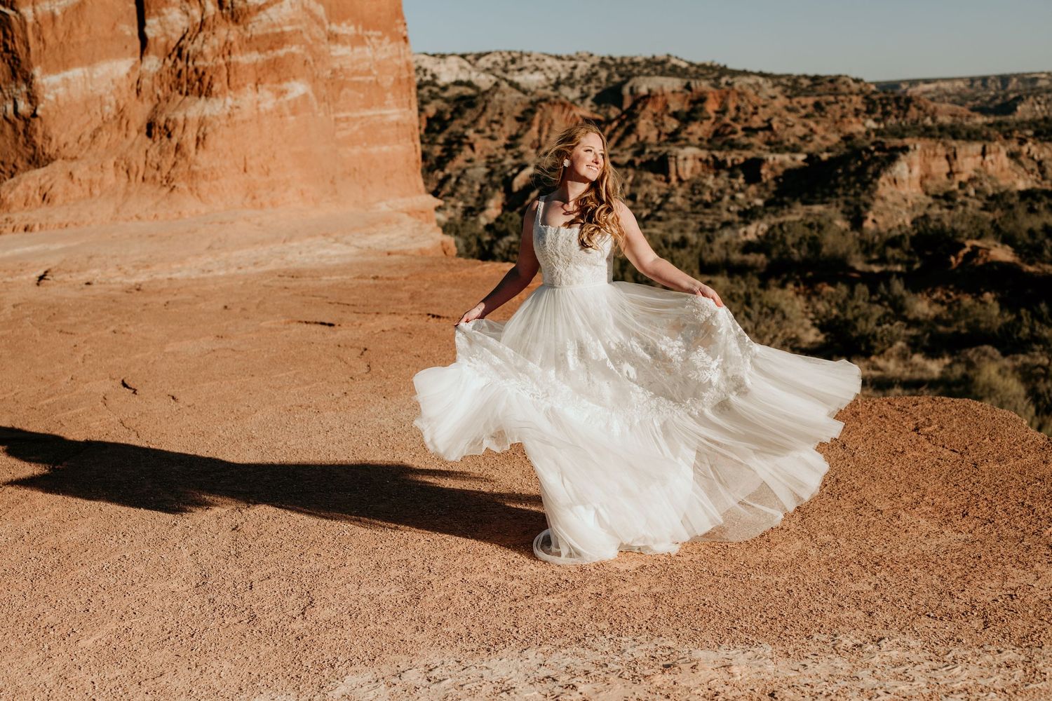 Flowing white wedding dress photographed against dramatic red rock formations in desert landscape.