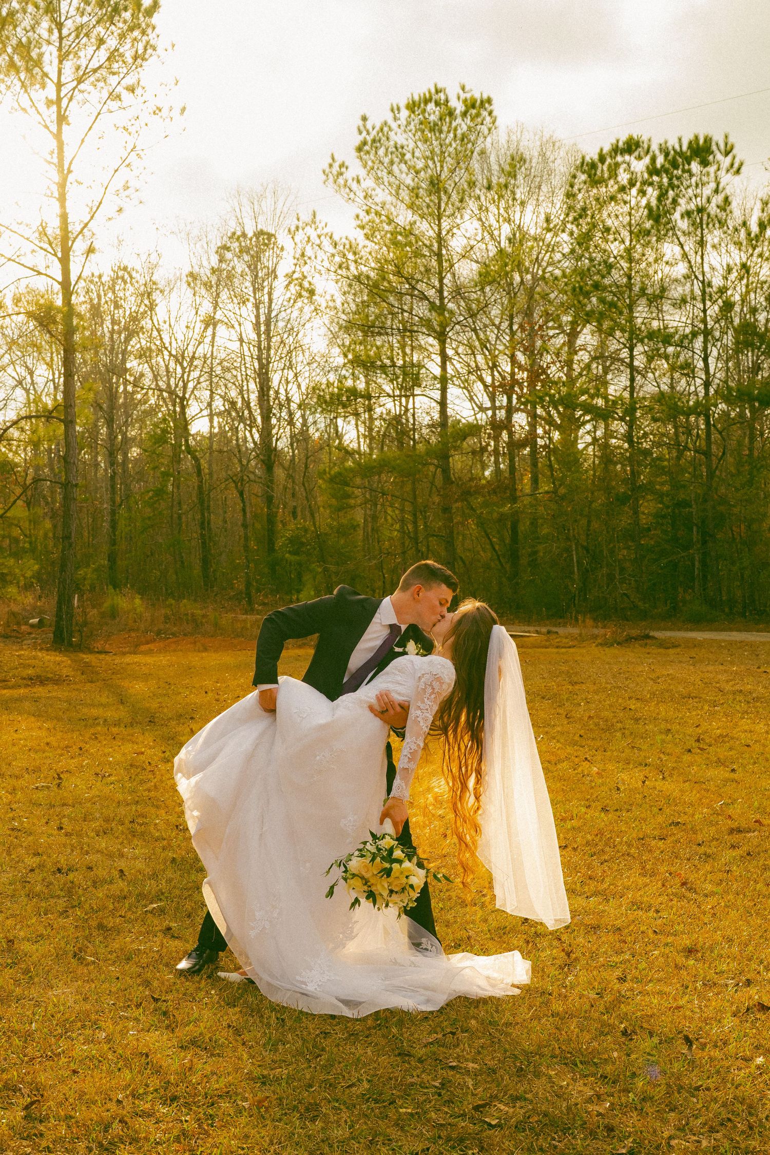 A romantic wedding couple shares a dip kiss in a golden autumn field surrounded by tall pine trees at sunset.