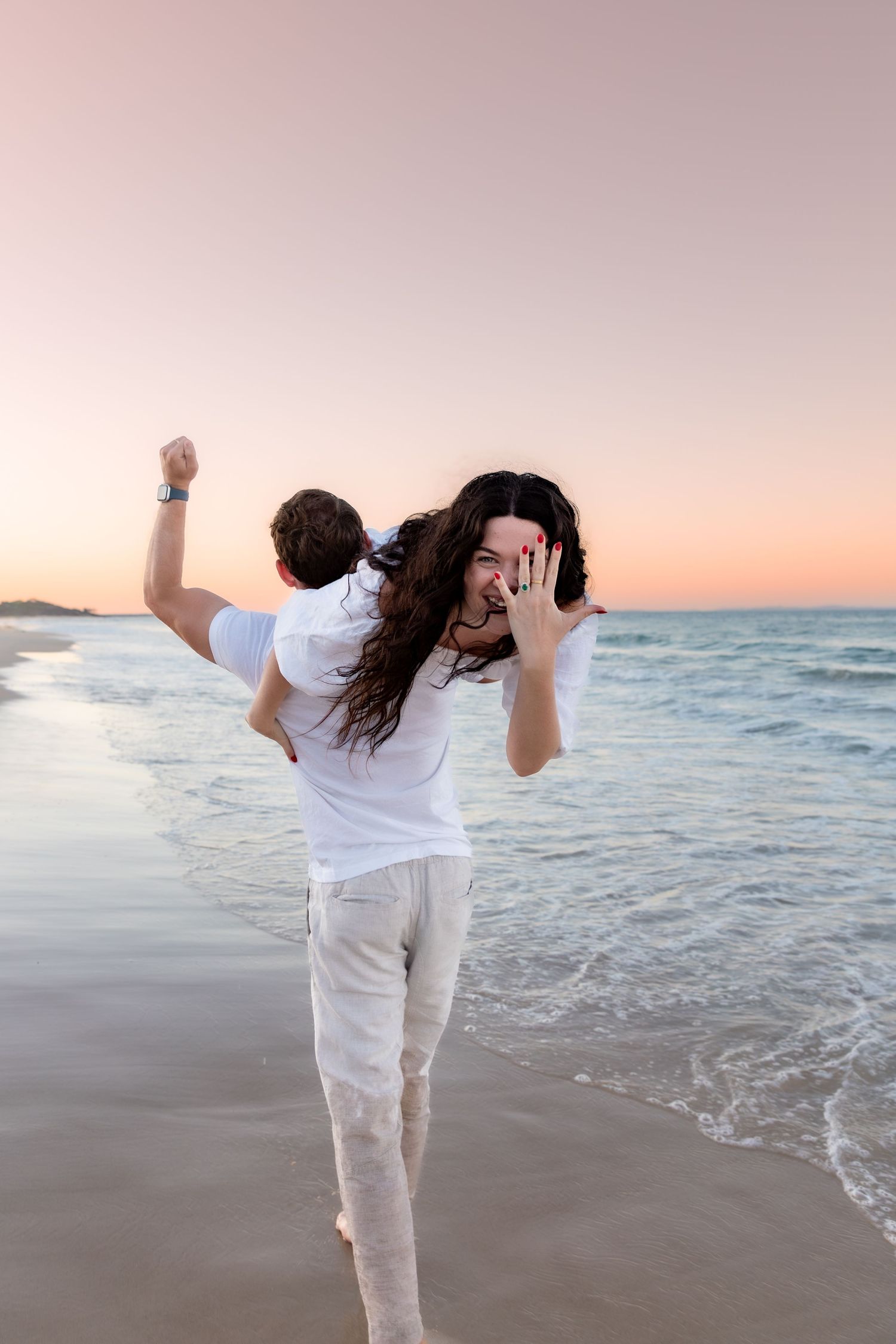 Two people in white outfits share a playful moment at the beach during sunset as waves roll in along the shore.